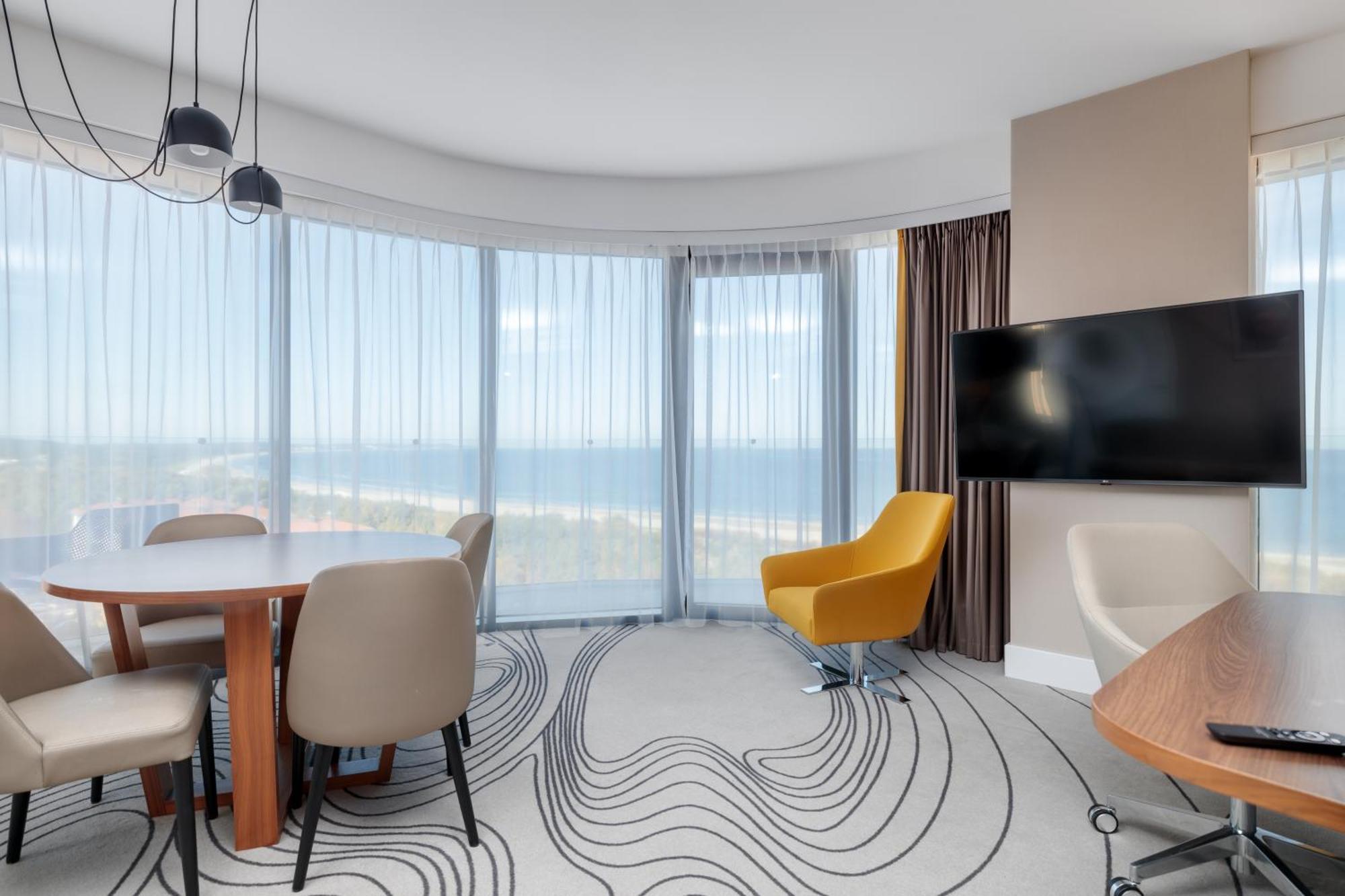 斯维诺乌伊希切丽笙布鲁酒店 外观 照片 The image shows a modern hotel room or suite with a bright, airy atmosphere. There are large windows that allow natural light to fill the space, providing a view of the ocean. The decor includes a round dining table with several chairs, and a stylish