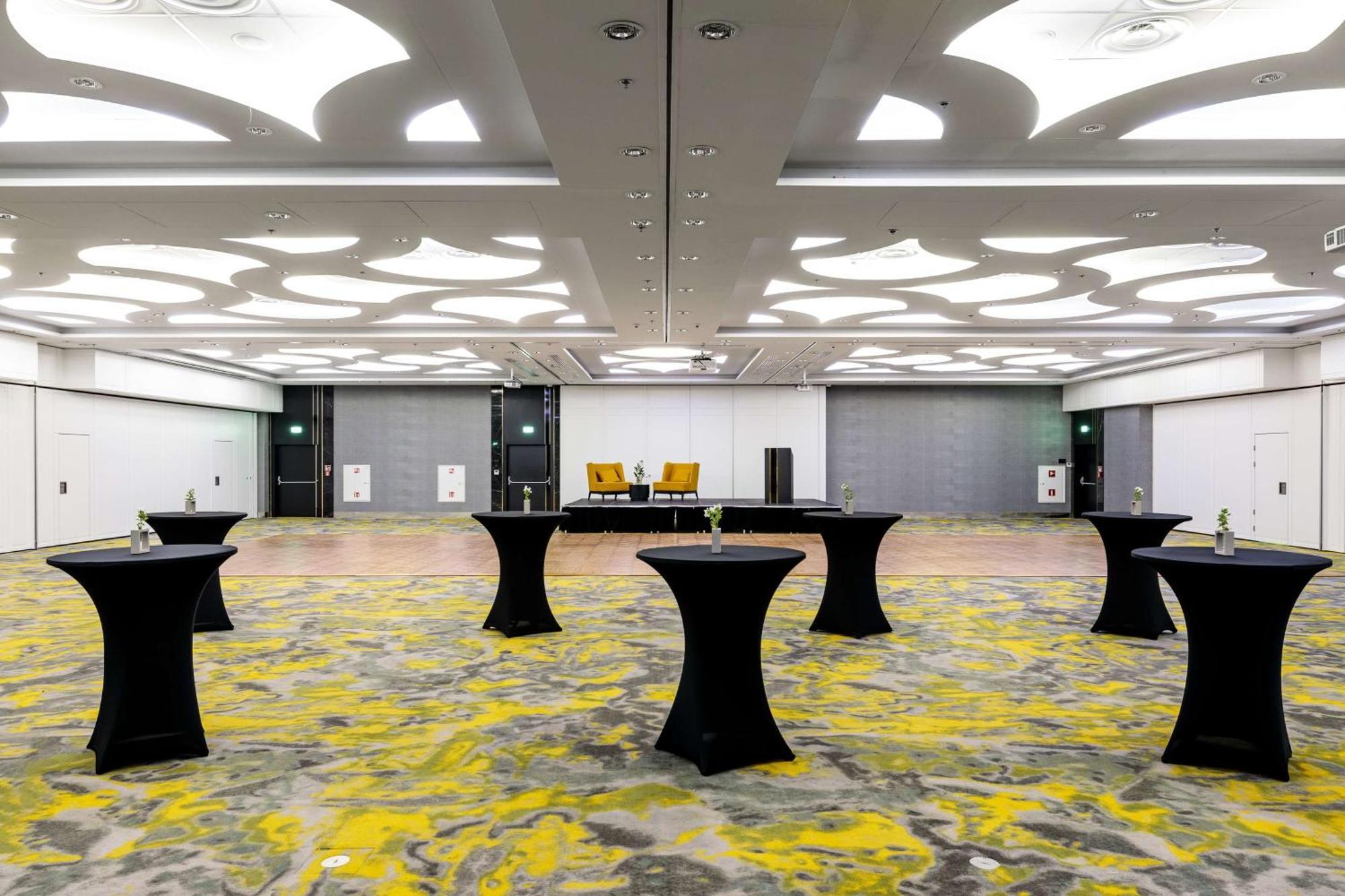 斯维诺乌伊希切丽笙布鲁酒店 外观 照片 The photo shows a spacious interior room set up for an event. The flooring features a patterned design in gray with yellow accents. There are several black tall tables scattered throughout the area, and in the background, there are two large yellow a