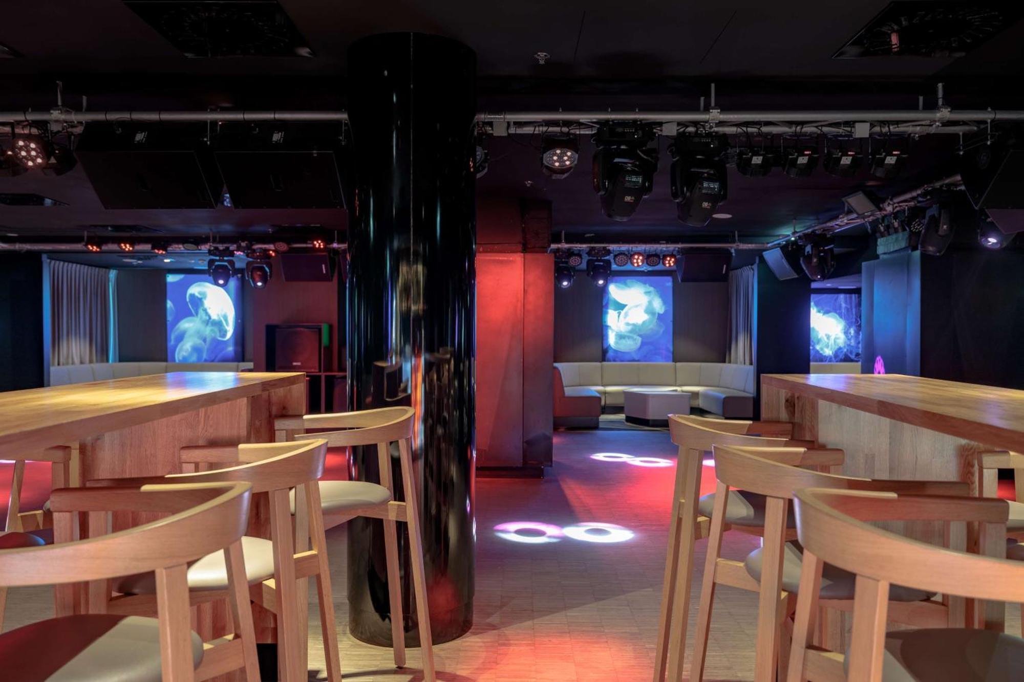 斯维诺乌伊希切丽笙布鲁酒店 外观 照片 The photo shows an interior of a modern venue, likely a bar or club. The space features a central black column and light-colored wooden bar stools arranged around a bar area. On the walls, there are large screens displaying vibrant visuals, possibly 